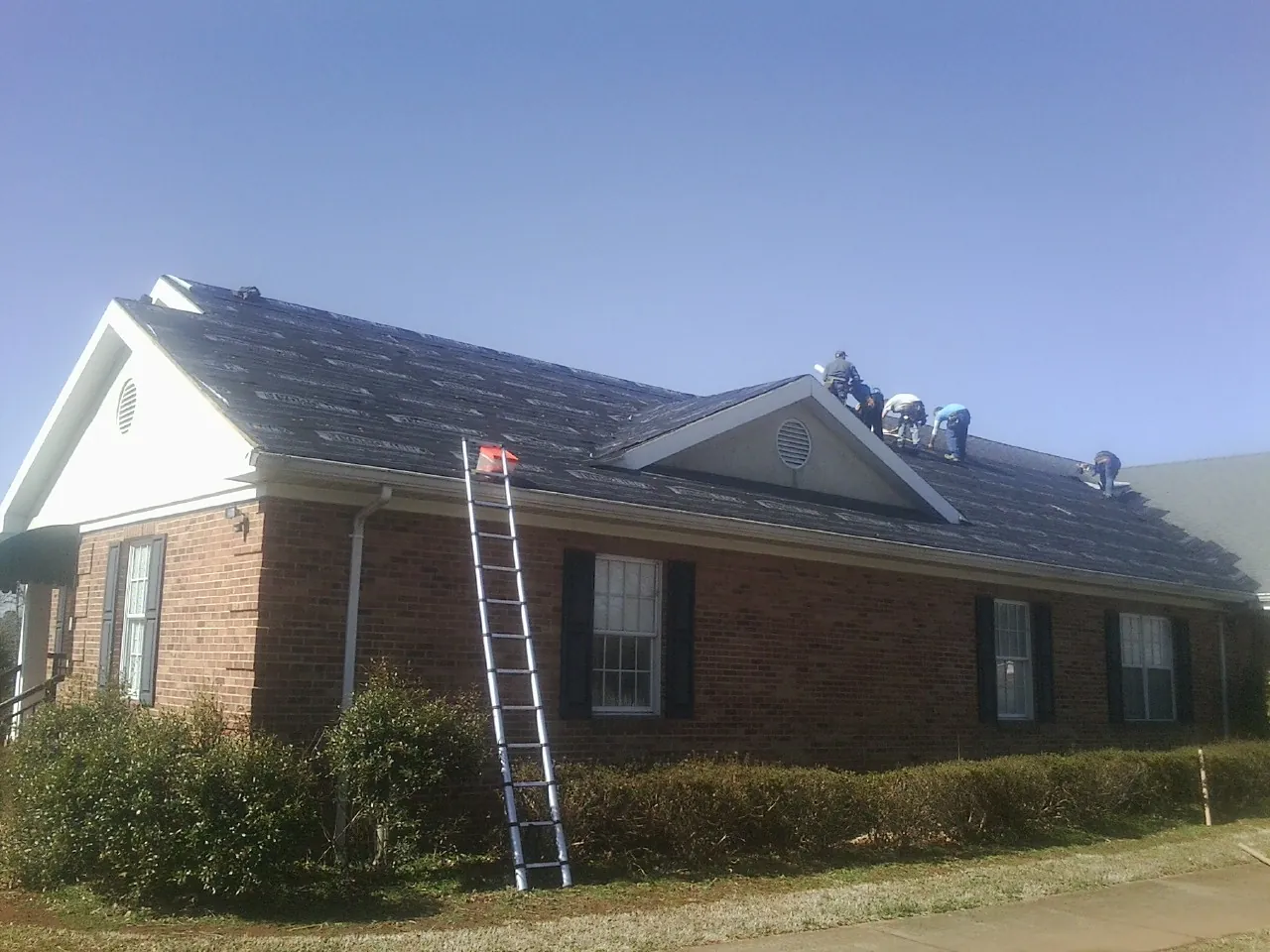 Crew tearing shingles off a brick ranch roof with a ladder set — Metal Roof Repair work by our crew in East Orange