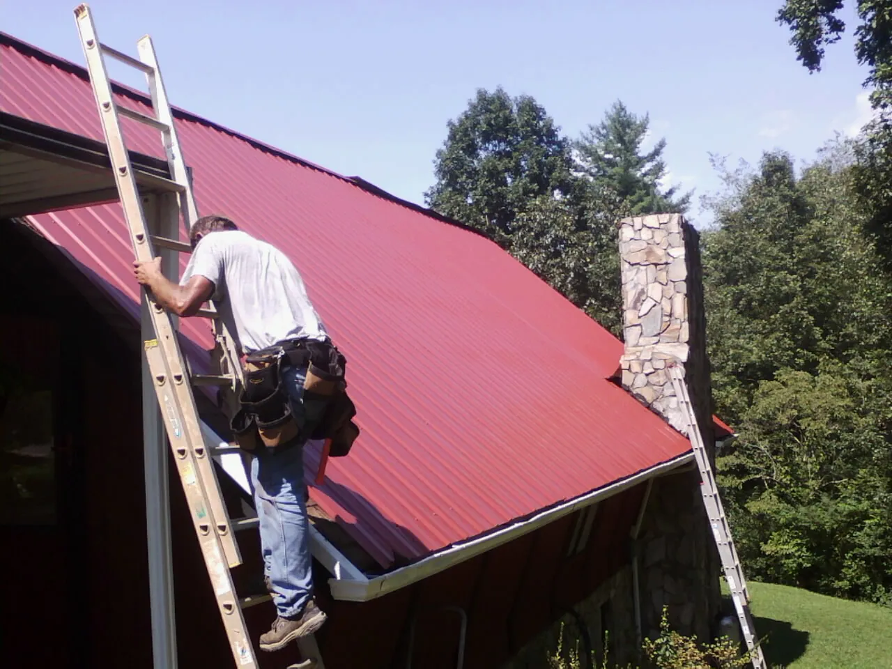 Roofer on a ladder beside a red metal roof and stone chimney — Soffit & Fascia Repair by Roofing Brothers in East Orange